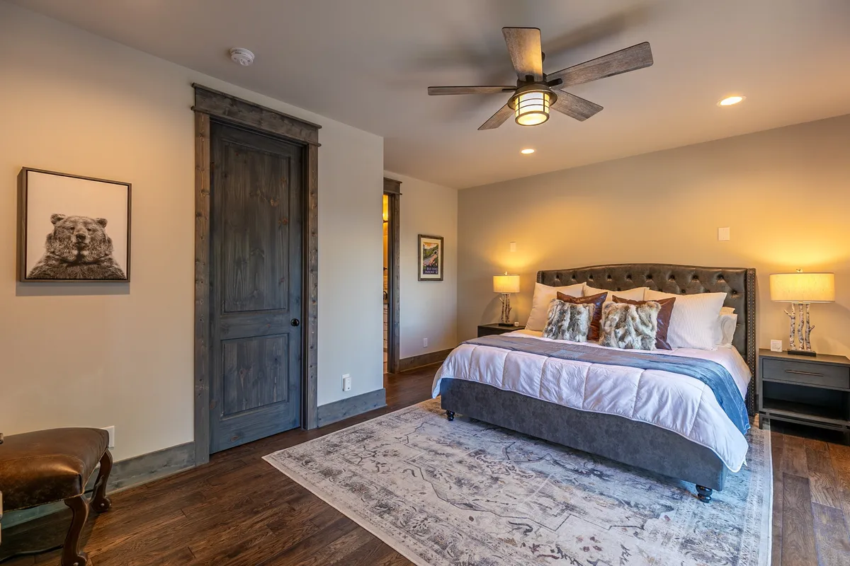 A tranquil bedroom featuring a comfortable bed with decorative pillows, soft natural light filtering in, rustic wooden details, and framed animal portrait above the headboard. Nightstands with lamps on either side provide a cozy ambiance, and the wooden door adds to the rustic-modern décor.
