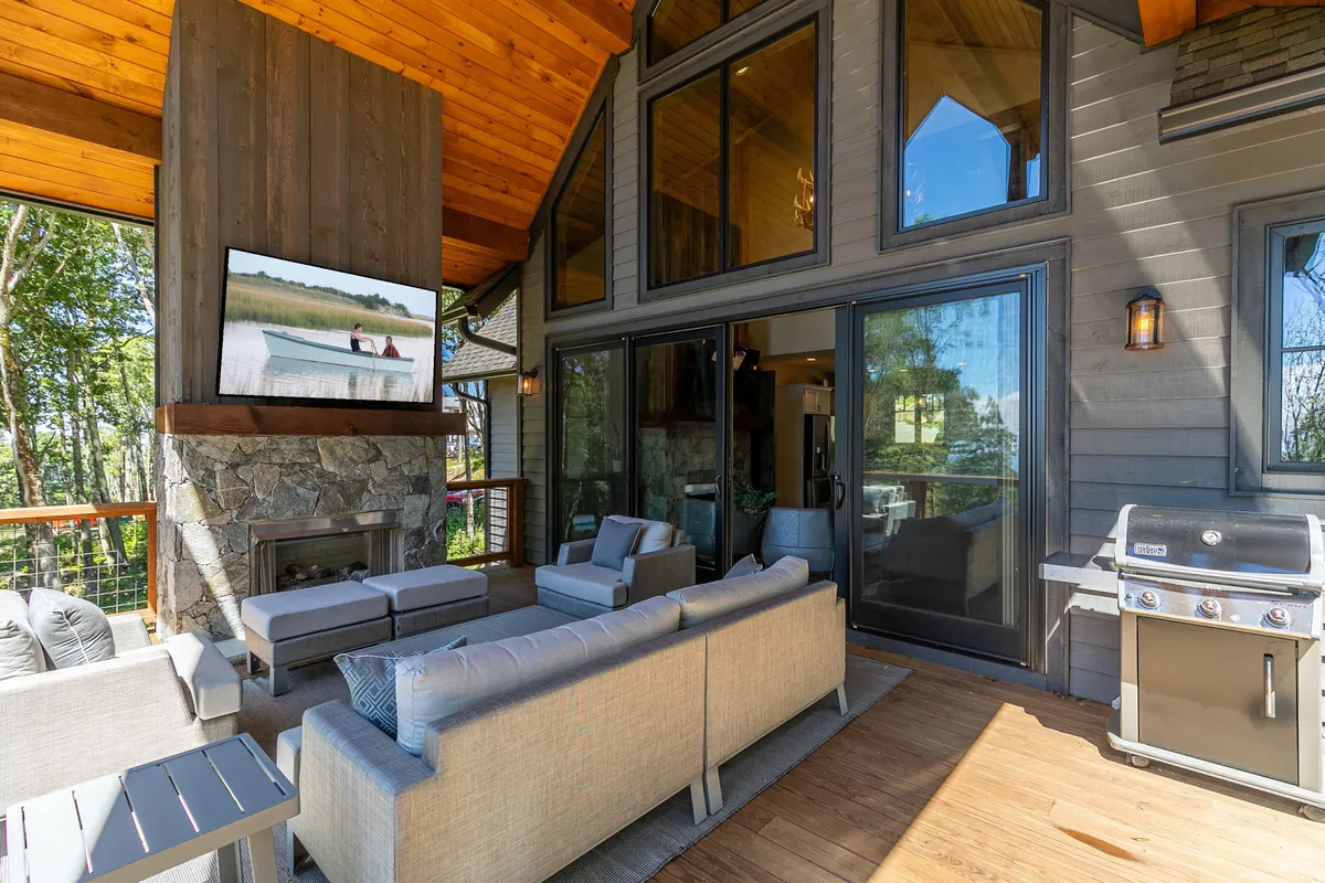 An inviting outdoor seating area with a fireplace, large windows, grill, and a wall-mounted TV. The scene is set up for guests to relax by the fire, watch sports or movies outdoors, dine al fresco, and enjoy social gatherings in comfort.