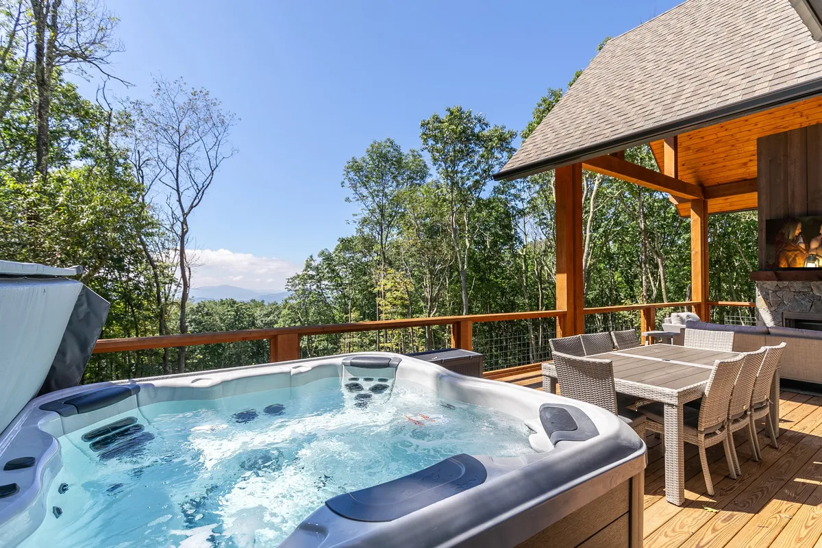 An inviting outdoor deck nestled among the trees, featuring a large hot tub and a dining set. The setting offers tranquil views, making it ideal for soaking under the stars or gathering for sunset drinks.