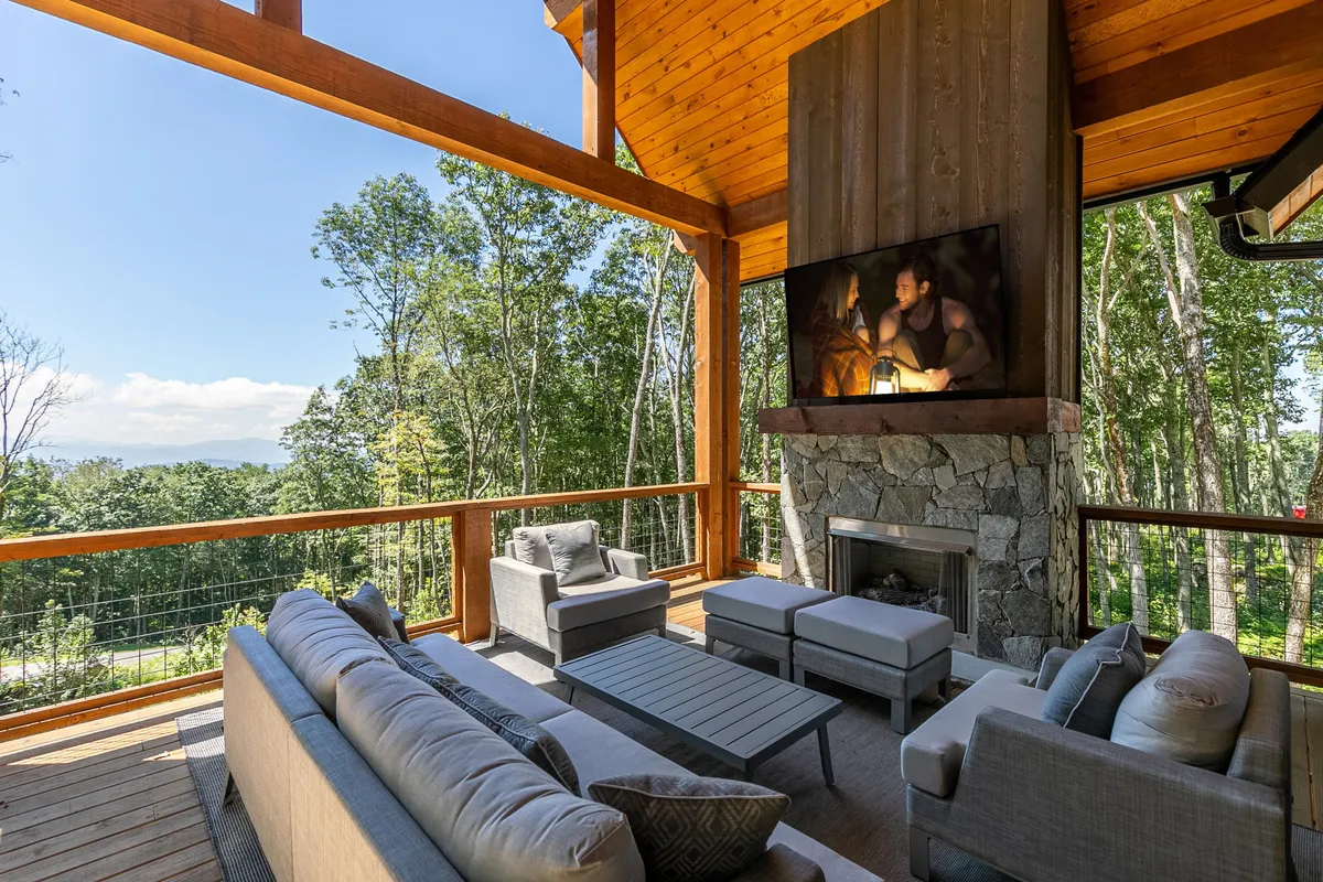 An inviting outdoor seating area with a fireplace, large TV, and forest views. The setup encourages relaxation, socializing, and entertainment—ideal for cozy gatherings after a day of outdoor activities, especially in cooler months. The presence of the fireplace and forested backdrop underscores the mountain resort ambiance.