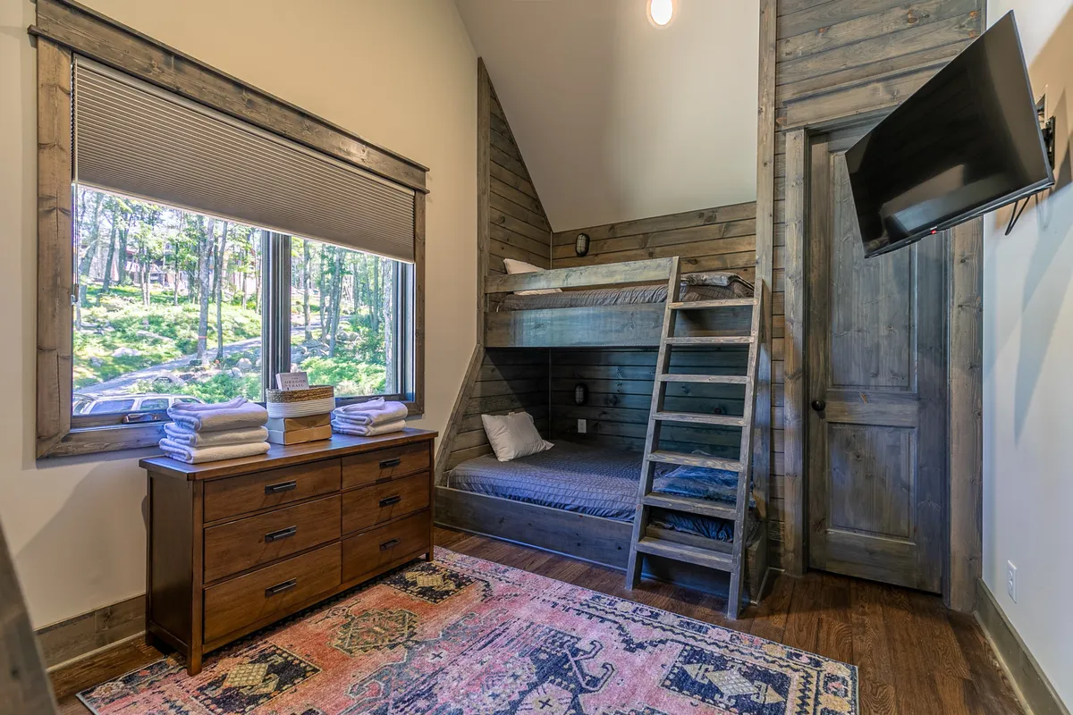 A cheerful bunk room featuring a whimsical bunk bed arrangement with twin beds and a queen, set against a bright window that brings in natural light. The space includes a cozy area rug, a playful dresser, and inviting decor ideal for children to read, play, or rest.