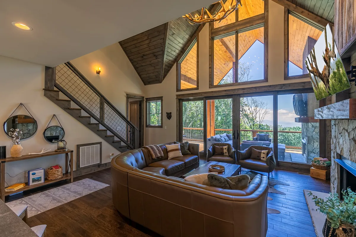 The image showcases a stunning cabin interior with soaring wooden beams, a cozy leather sofa facing a grand stone fireplace, and floor-to-ceiling windows framing sweeping views of the Blue Ridge Mountains. Warm natural light pours in, creating an inviting space perfect for relaxation or gathering with loved ones.