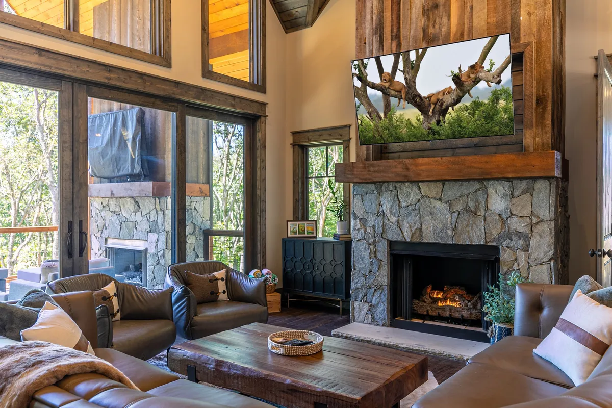 A warm, welcoming living room with a fireplace as the focal point, flanked by large windows that frame serene natural views. The comfortable seating and entertainment setup suggest a perfect spot for cozy evenings, movie nights, or enjoying the scenery.