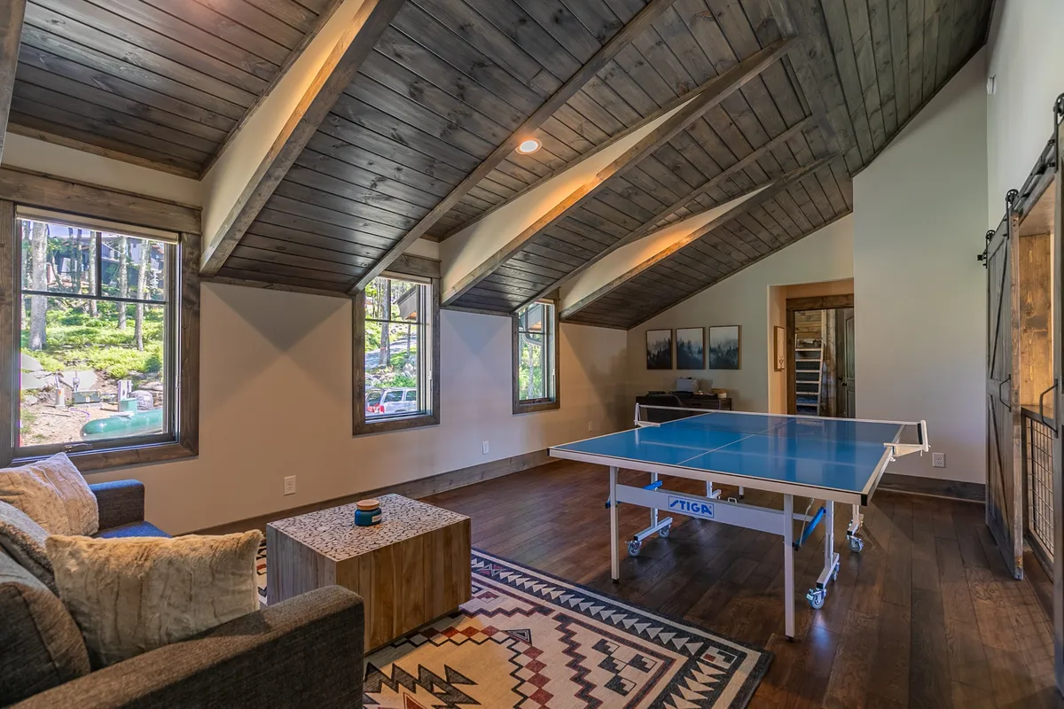 A casual recreation room with a ping pong table, a cozy sofa, a wooden coffee table, and windows that hint at nature views beyond. The space invites socializing and unwinding after a day outside.