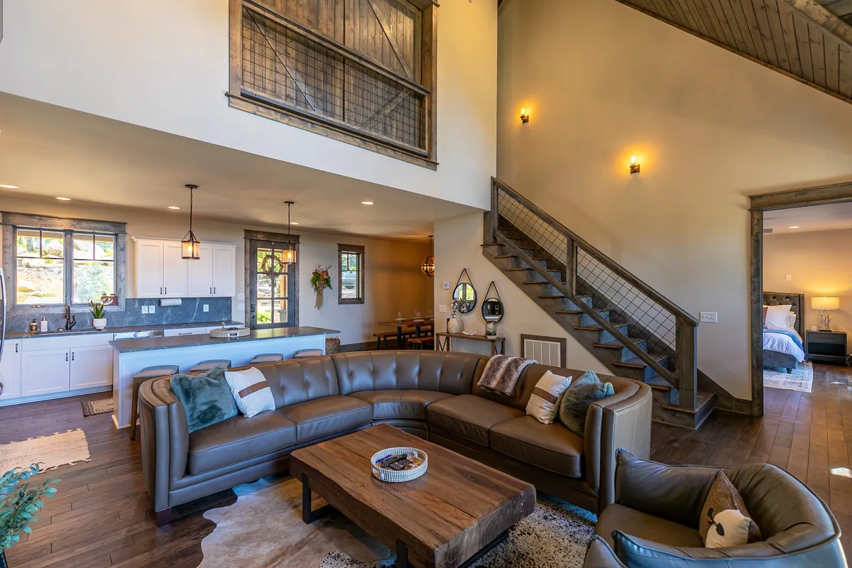 A spacious open-concept living area featuring a large sectional sofa, a modern wooden coffee table, a staircase, and an open kitchen. Natural light pours in, highlighting the cozy, inviting atmosphere ideal for both relaxation and entertaining. The setting hints at luxury with its upscale furnishings and thoughtful layout.