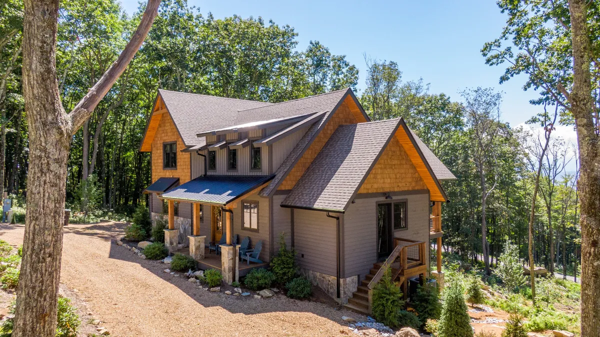 A peaceful exterior view of the property with a wooden façade, stone pillars, and surrounding trees. The gravel pathway and porch suggest a welcoming entrance and a space to enjoy sunsets or campfire nights, capturing the retreat's tranquil mountain setting.