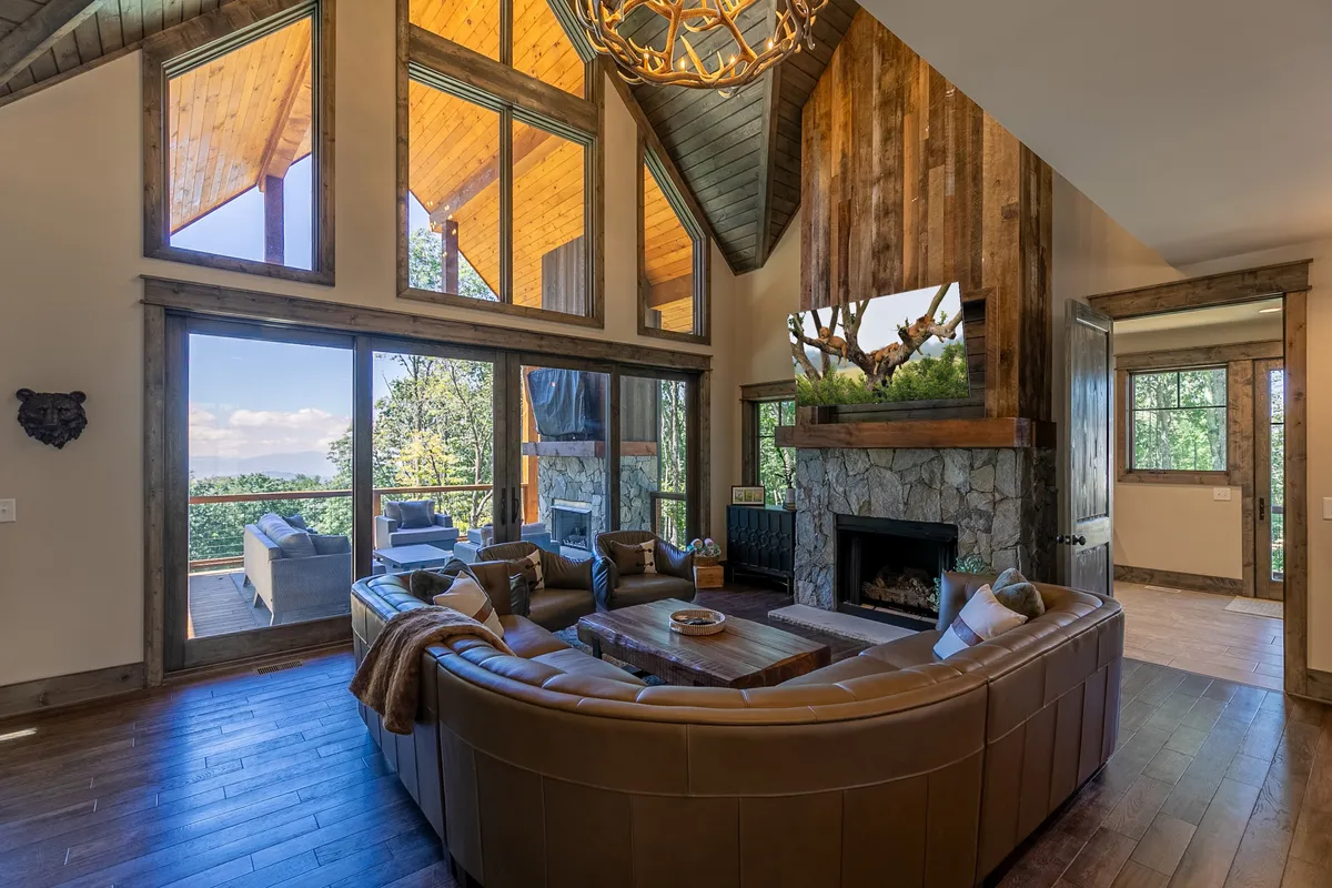A spacious living room featuring large windows that frame stunning views of the Blue Ridge Mountains. The space includes a chic leather sofa, a statement stone fireplace, and an antler chandelier, creating a warm, inviting atmosphere perfect for relaxation and intimate gatherings.