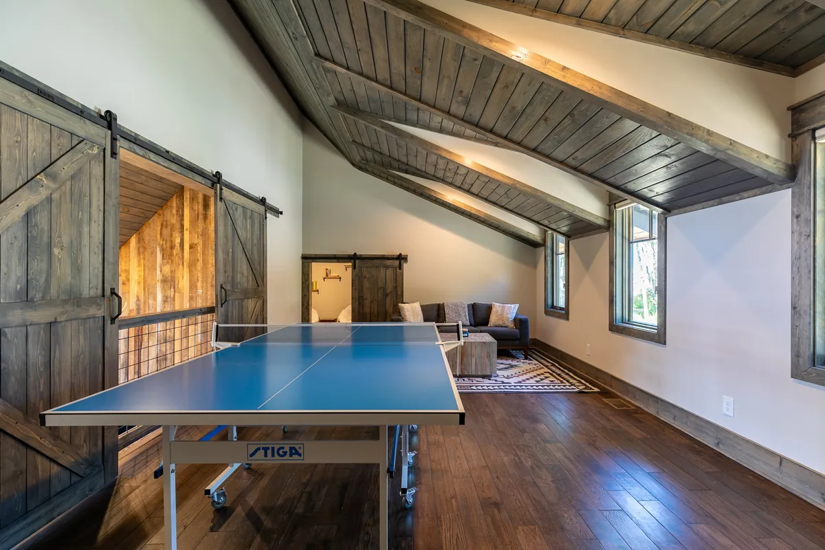 A vibrant recreational space centered around a ping pong table, with a comfortable sofa, rustic wooden beams, and sliding barn doors creating an energetic yet relaxed ambiance. Ideal for friendly competitions and group games.